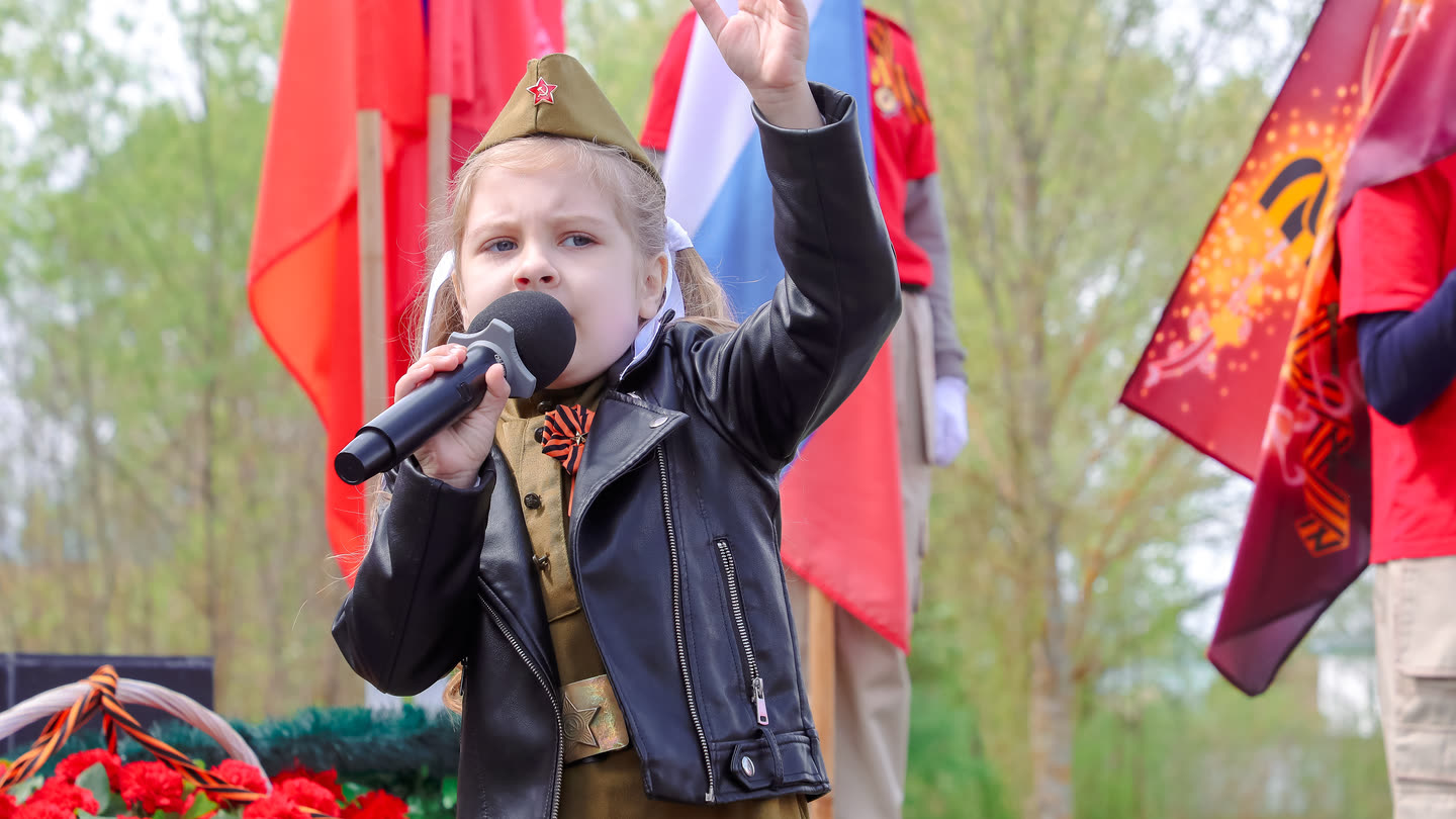 Алина Бер поёт в микрофон на памятном мероприятии 9 Мая: пилотка, гимнастёрка и чёрная косуха, за спиной флаги и красные гвоздики